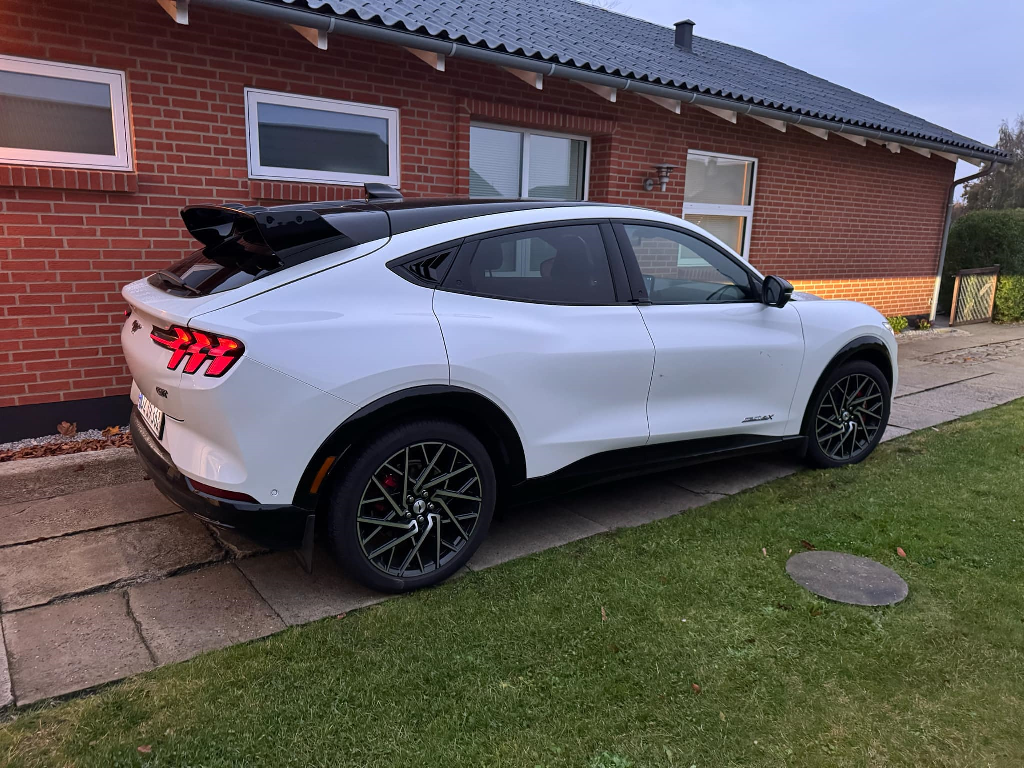 White Ford Mustang Mach-E with black Rally Wing Spoiler installed on rear hatch, parked beside a brick house.