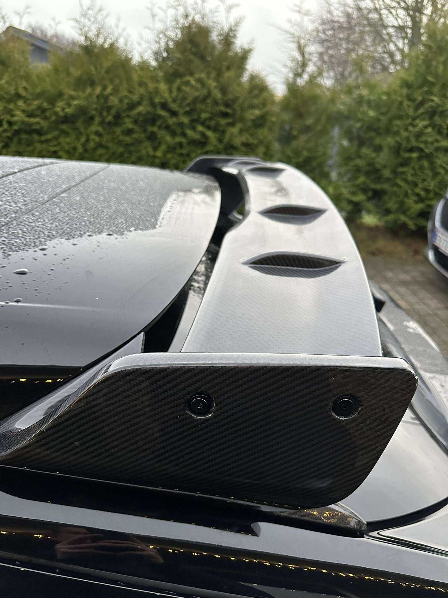 Close-up of a carbon fiber Rally Wing Spoiler installed on a black Ford Mustang Mach-E, showcasing its aerodynamic design and glossy finish with raindrops on the surface.